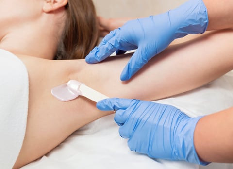 Armpit waxing procedure in a beauty salon. Master in gloves applies wax with a wooden spatula to a woman's armpit to remove hair