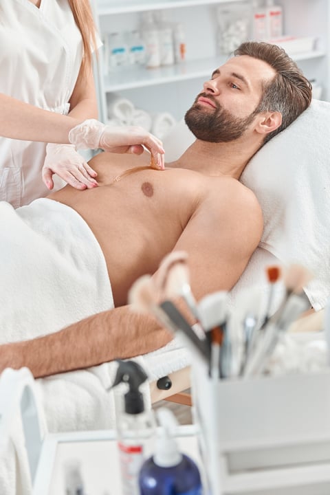 Woman beautician put depilatory wax to chest of young man for hair removal. Depilation with wax.