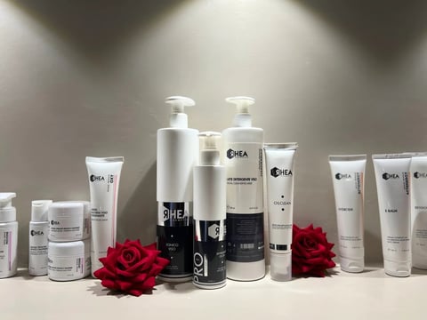 Collection of ЯHEA skincare and cosmetic products displayed with red roses, including pump bottles, tubes, and jars against a gray background